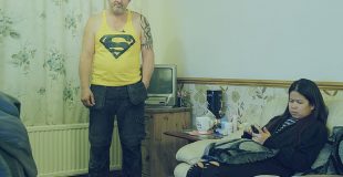 На фото в комнате стоит полноватый мужчина в желтой майке со значком супермена. На диване рядом сидит женщина и смотрит в телефон.