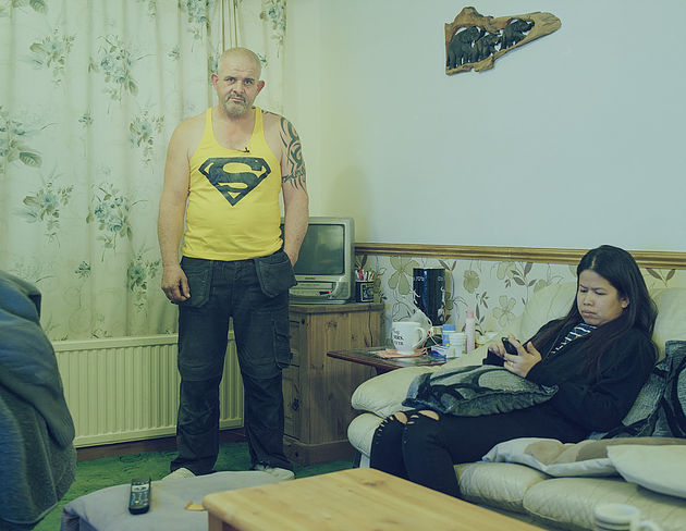 На фото в комнате стоит полноватый мужчина в желтой майке со значком супермена. На диване рядом сидит женщина и смотрит в телефон.