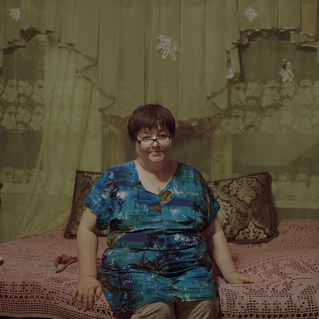 На фото женщина в халате и очках, она сидит на диване. На фоне занавески с рюшками.