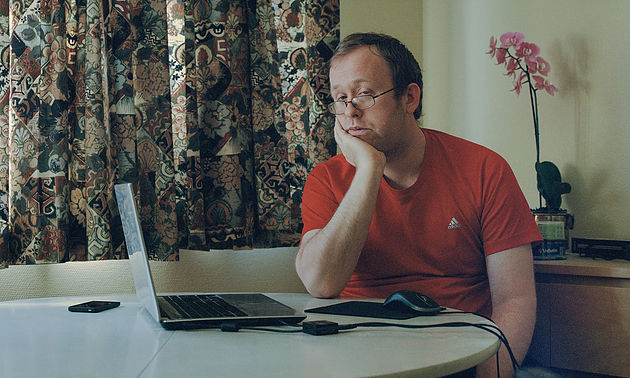 На фото мужчина, он сидит за столом подперев подбородок рукой и смотрит в ноутбук.